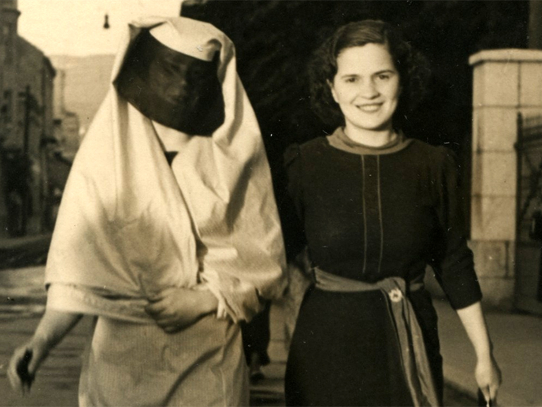 A woman wearing a dark urban dress is walking down the street with a Muslim woman wearing a veiled robe and a black veil covering her face.